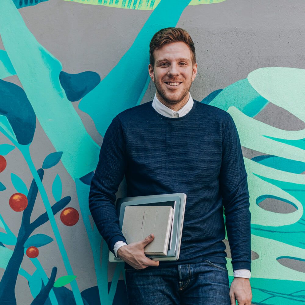 Homme tenant un dossier de notes et un ordinateur portable fermé, adossé à un mur coloré